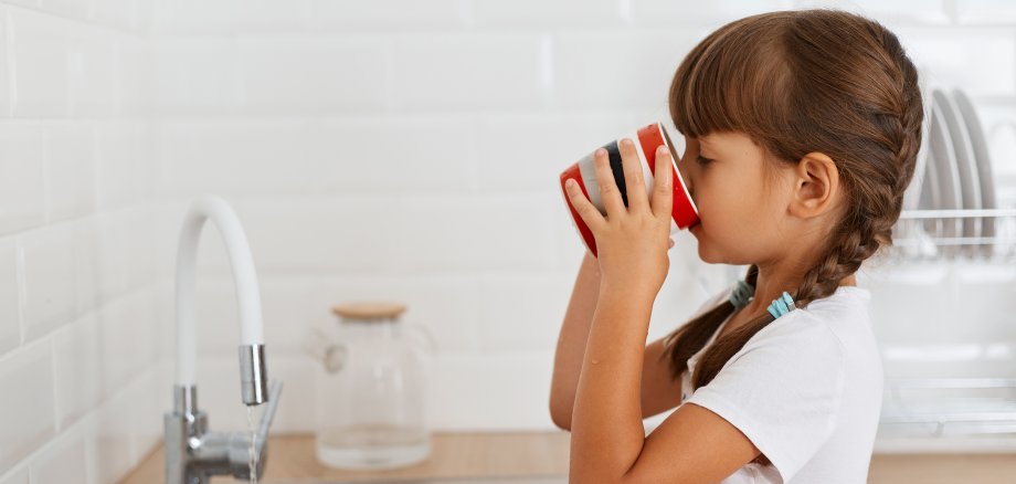 Side view portrait of dark haired little girl with pigtails wearing white t shirt holding cup and standing near the kitchen faucet, drinking clean water, feels thirsty.